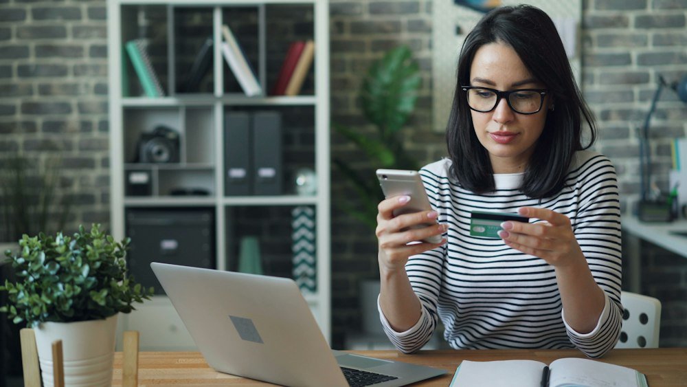 Mulher com cartão na mão conferindo no celular se o site é confiável antes de pagar